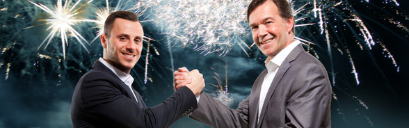 Two men in suits shaking hands in front of a fireworks display
